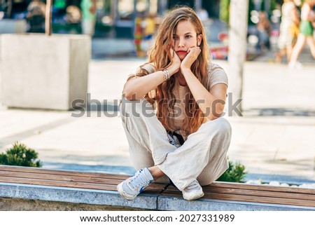 Pensive young woman with long hair resting on the street. Full-length view of sad university student sitting outside waiting for her colleague outdoor.