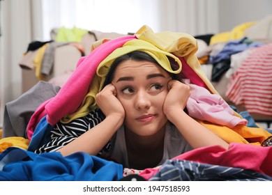 Pensive Young Woman With Lots Of Clothes On Floor In Room. Fast Fashion