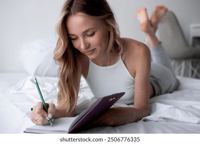 Pensive young woman lying on bed with serious expression writing on journal. copy space