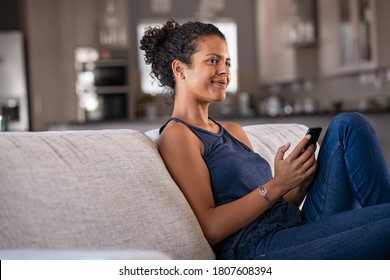 Pensive Young Woman Sitting On Couch And Holding Smartphone While Looking Away. Smiling African Woman Daydreaming And Holding Mobile Phone With Copy Space. Black Girl Thinking While Using Smart Phone.