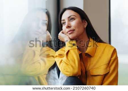 Pensive young woman staring out of a window with a faraway expression reflected in the glass