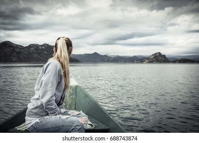 Pensive Young Woman Tourist Looking At Beautiful Landscape On   Bow Of Boat Floating On Water  Towards Shore In Overcast Day With Dramatic Sky  