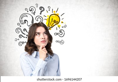 Pensive Young Woman With Wavy Dark Hair Standing Near Concrete Wall With Question Marks And Lightbulb Drawn On It. Concept Of Bright Idea, Education And Choice. Mock Up