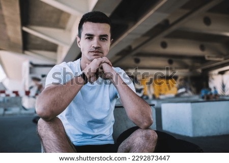 Pensive youthful male wearing t shirt shorts and wristwatch sitting on ground under bridge during daylight and looking at camera