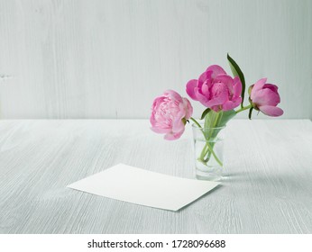 Peonies bouquet in clear glass vase. Clean scandinavian style decoration. Empty invitation card mock-up on white wooden table.