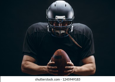 People, Achievement And Sport Concept. Athletic American Football Player In Black Helmet And Jersey Posing With A Ball Isolated On Black Background