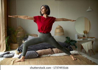 People, activity, health and vitality concept. Stylish barefoot young woman exercising at home, doing vinyasa flow yoga in her bedroom, standing on carpet in virabhadrasana or warrior II pose - Powered by Shutterstock