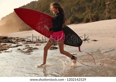 People and adventure concept. Active brave surfboarder runs quickly as notices big wave, wants to hit it, being in good mood, tries new surfboard, poses against cliff and sandy beach background