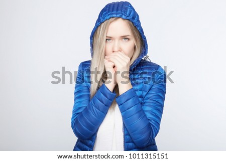 People, adventure, traveling concept. Young blond lady freezing, wearing thin blue demi-season jacket with hood on head, looking at camera with deep blue eyes, trying to warm herself with breath. 