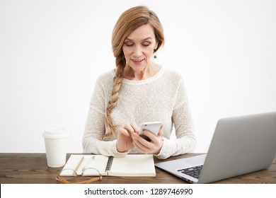 People, Age, Electronic Gadgets And Online Communication Concept. Positive Attractive Mature Sixty Year Old Female Sitting At Her Workplace In Front Of Open Laptop, Texting Sms Using Mobile Phone