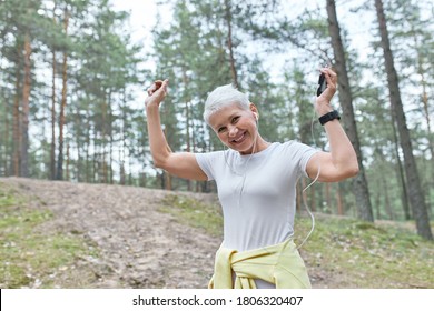 People, Age, Fun And Active Lifestyle Concept. Happy Middle Aged Woman Listening To Running Playlist Using Smart Phone. Healthy Fit Mature Female In Earphones Jogging Or Dancing In Park, Having Fun