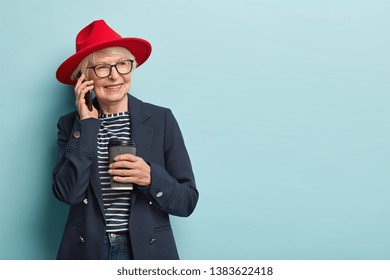 People, Age And Leisure Concept. Happy Old Lady Enjoys Spare Time, Has Telephone Conversation, Drinks Coffee To Go, Wears Red Headgear And Formal Coat, Looks Aside, Models Over Blue Wall, Free Space