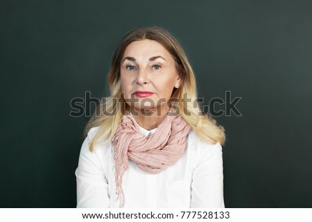 People, age and lifestyle concept. Picture of beautiful Caucasian retired woman with loose blonde colored hair, blue eyes and wrinkles posing in studio, wearing stylish mustard scarf over white blouse