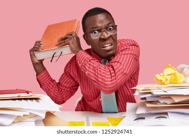 People, anger, threat concept. Irritated male teacher holds big encyclopedia, going to revenge with someone, feels annoyed with constant studying, wears pink shirt, has no spare time for rest