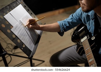 people, art and entertainment concept - man with guitar writing notes to music book at studio