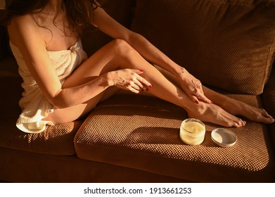 People, Beauty, Depilation, Epilation, Body Care Concept. Beautiful Woman Bare Legs Sitting On Couch At Home On Bright Sunny Day. Young Lady In Towel After Shower Applies Moisturizer Long Tanned Legs