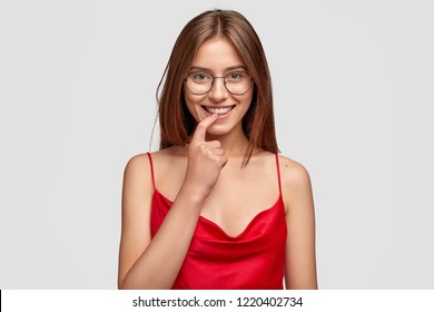 People, Beauty And Happy Emotions Concept. Glad Lovely Woman Bites Finger, Looks Joyfully At Camera, Recieves Compliment From Stranger, Wears Round Big Spectacles And Red Dress, Models In Studio