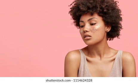 People, beauty and make up concept. Adorable pretty female with dark curly hair, has glitter on face, looks thoughtfully down, stands against pink background, has Afro hairstyle and pure healthy skin