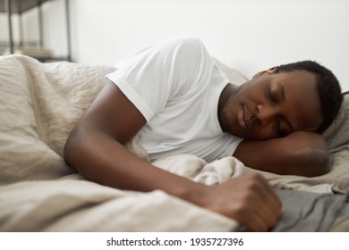 People, bedding, bedtime, rest and relaxation concept. Indoor shot of handsome African American male wearing white t-shirt lying in bed, sleeping peacefully, having pleasant good dream