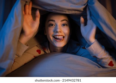 people, bedtime and rest concept - happy teenage girl lying under blanket in bed with her tongue stuck out at home at night