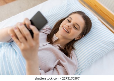 people, bedtime and rest concept - happy smiling teenage girl with smartphone lying in bed at home