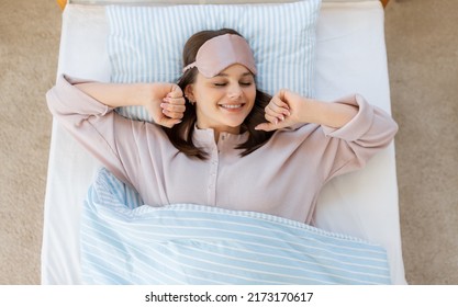 people, bedtime and rest concept - happy smiling teenage girl in eye sleeping mask stretching in bed at home in morning
