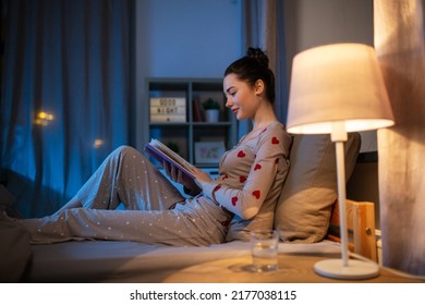 people, bedtime and rest concept - happy teenage girl reading book in bed at home at night