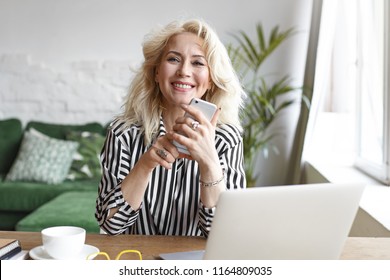 People, Business, Age And Modern Gadgets Concept. Cheerful Successful Sixty Year Old Blonde Self-employed Woman Working From Home, Using Generic Electronic Devices, Enjoying Online Cmmunication