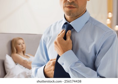 People, Business, Relations And Problems Concept - Close Up Of Man In Shirt Dressing Up And Adjusting Tie On Neck Over Woman In Bed Background