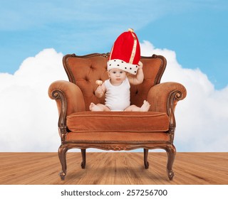 People, Childhood And Royalty Concept - Happy Baby Boy In Royal Hat With Lollipop Sitting On Chair Over Blue Sky And Cloud Background