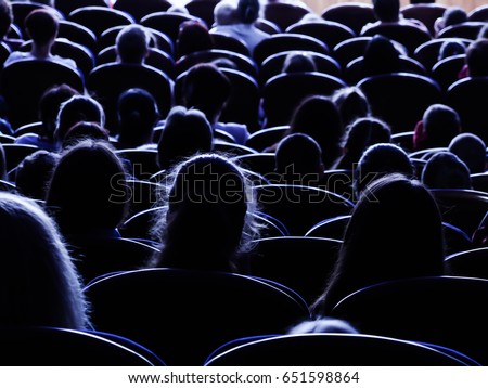People, children, adults, parents in the theater watching the performance. People in the auditorium looking at the stage. Shooting from the back