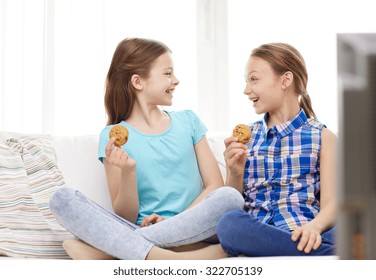 people, children, television, friends and friendship concept - two happy little girls watching tv and eating cookies at home