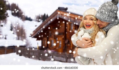 People, Christmas, Holidays And Season Concept - Happy Family Couple In Winter Clothes Hugging Over Wooden Country House And Snow Background