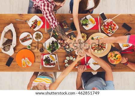 People clink glasses, saying cheers, eat healthy meals at party dinner table. Friends celebrate with organic food, ratatoille and corn barbecue on wooden table top view. Woman pass dish plate to man