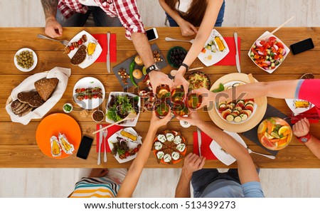 People clink glasses, saying cheers, eat healthy meals at party dinner table. Friends celebrate with organic food, ratatoille and corn barbecue on wooden table top view.