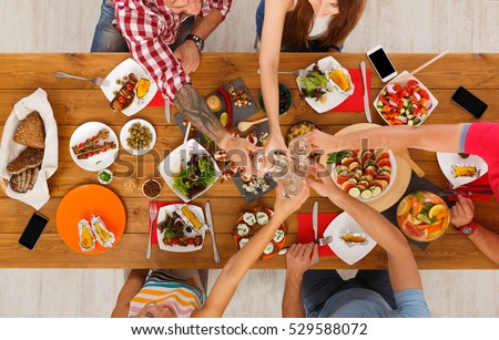 People clink glasses, saying cheers, eat healthy meals at party dinner table. Friends celebrate with organic food, ratatoille and corn barbecue on wooden table top view. Woman pass dish plate to man