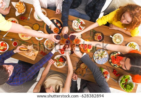 People clink glasses, saying cheers, eat healthy meals at party dinner table. Friends celebrate with organic food, ratatoille and corn barbecue on wooden table top view.