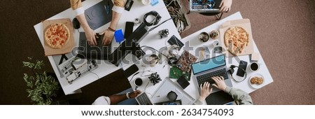 People collaborating on technology projects at office workspace, surrounding by laptops, pizza boxes, electronic components, and coffee cups. Hands visible, working on various tasks