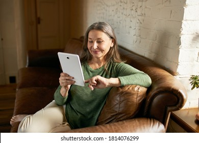 People, communication, electronic gadgets and devices. Beautiful woman pensioner relaxing on comfortable leather sofa with digital tablet, playing video games or chatting online via social networks - Powered by Shutterstock