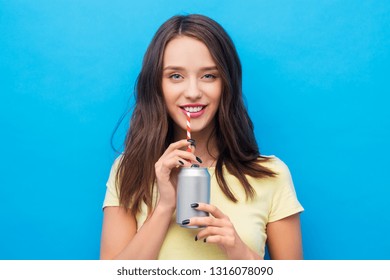 People Concept - Smiling Young Woman Or Teenage Girl In Yellow T-shirt Drinking Soda From Can Through Paper Straw Over Bright Blue Background