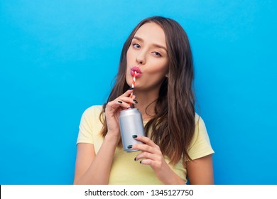 People Concept - Smiling Young Woman Or Teenage Girl In Yellow T-shirt Drinking Soda From Can Through Paper Straw Over Bright Blue Background