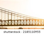 People cross a suspension bridge at sunset. Silhouette profile shot of bridge with low tree-line and bridge in background. 