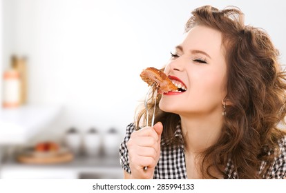 People, Diet, Culinary And Food Concept - Hungry Young Woman Eating Meat On Fork Over Kitchen Background