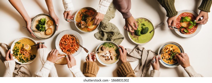 People Eating Autumn And Winter Creamy Vegan Soups, Fall And Winter Vegetarian Food Menu. Flat-lay Of Peoples Hands With Homemade Soup In Plates And Bread Slices Over White Table Background, Top View