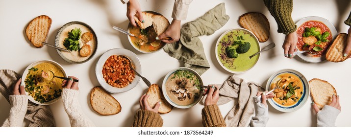 People Eating Autumn And Winter Creamy Vegan Soups, Fall And Winter Vegetarian Food Menu. Flat-lay Of Peoples Hands Eating Homemade Soup With Fresh Bread Over White Table Background, Top View
