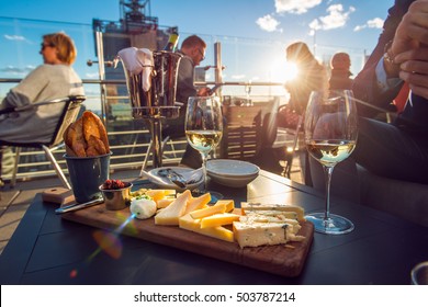People eating cheese and drinking wine at rooftop restaurant at sunset time. Restaurant table served with cheese plate, bread and white vine full of visitors. - Shutterstock ID 503787214