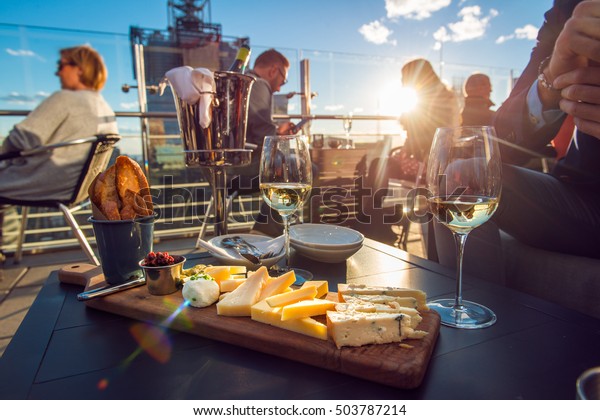 People eating cheese and drinking\
wine at rooftop restaurant at sunset time. Restaurant table served\
with cheese plate, bread and white vine full of\
visitors.