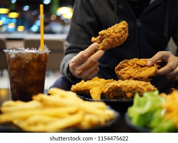 People Eating Fast Food Concept Hand Holding Deep Fried Chicken In The Restaurant With Blurred Foreground Of French Fries, Vegetable Salad, Carbonated Soft Drink And Bokeh Lights Background.