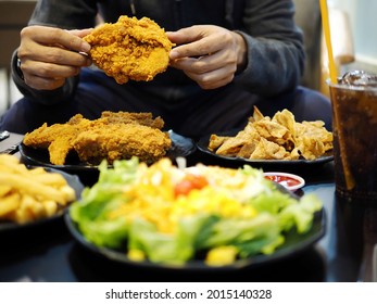 People Eating Fast Food Concept Hand Holding Deep Fried Chicken In The Restaurant With Blurred Foreground Of French Fries, Vegetable Salad, Carbonated Soft Drink And Bokeh Lights Background.