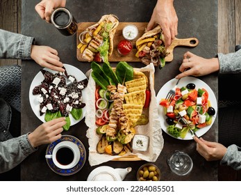 People eating Greek food. Greek cuisine assortment on dark tabletop, top down view. Hands with gyros, greek salad, souvlaki - selected of greek cuisine